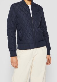 Navy quilted bomber jacket with a zip closure, ribbed cuffs, and two side pockets. Soft, smooth texture with diamond pattern detailing.