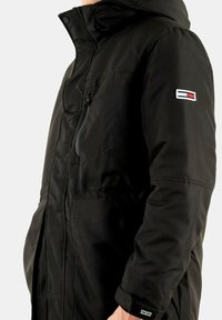 Giacca esterna nera con cappuccio, zip frontale e tasche laterali. Include una toppa con logo con accenti rossi, bianchi e blu sulla manica.