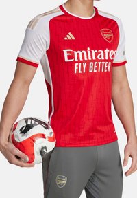 Red Adidas Arsenal jersey with white accents, zigzag pattern, gold detailing, and white lettering. Gray pants with golden logo. Individual holding a football.