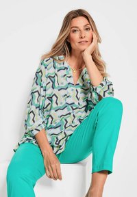 Bluse mit geometrischem Muster in Grüntönen, Blau und Schwarz; leichtes Material; V-Ausschnitt; kombiniert mit einfarbigen türkisfarbenen Hosen.
