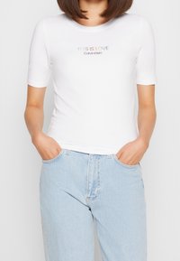 Osoba nosící bílý krátký rukáv top od Calvina Kleina s nápisem "TOTO JE LÁSKA" v pastelových barvách a světle modré džíny s rukama v kapsách.