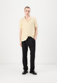 Kurzarm-Hemd mit Knopfleiste aus hellgelbem strukturiertem Stoff, kombiniert mit schwarzen gefalteten Hosen und schwarzen Schuhen. Minimalistisches Design.