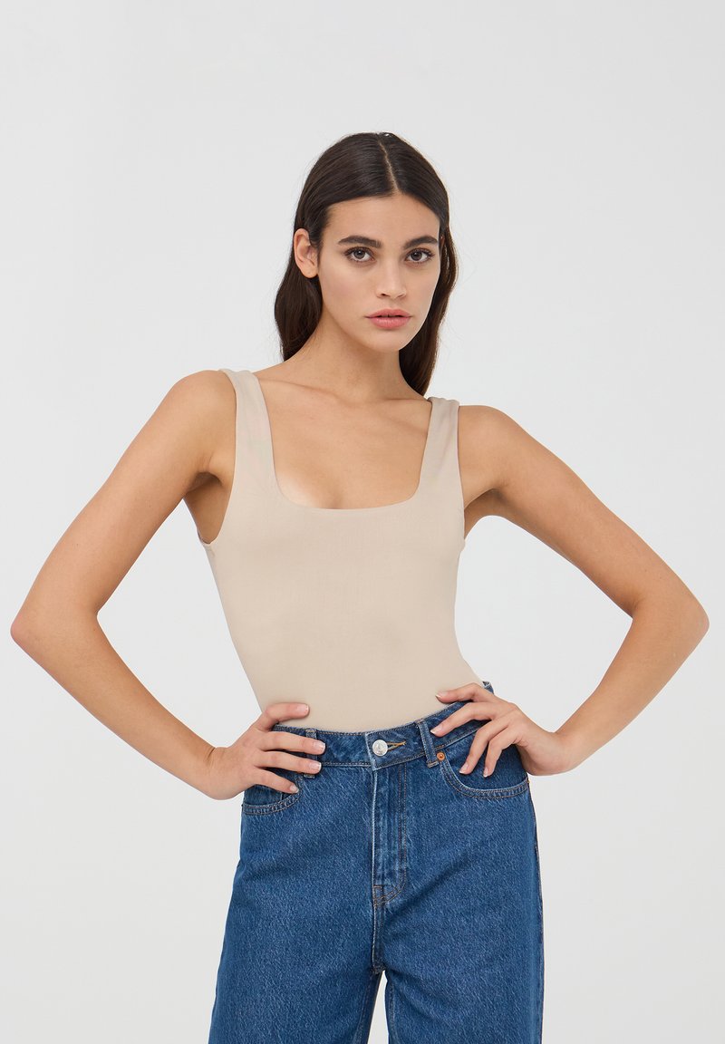 Body beige senza maniche con scollo quadrato, abbinato a jeans in denim blu a vita alta. Il tessuto appare liscio e aderente.