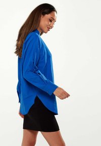 Blue long-sleeve shirt with a button-down collar, featuring a relaxed fit, side slits, and soft texture, paired with a black mini skirt.