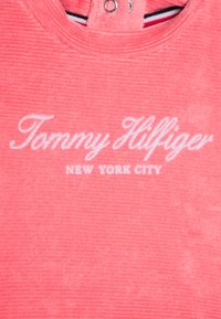 Różowo-koralowa ryflowana tkanina z haftowanym białym napisem "Tommy Hilfiger" pisanym kursywą oraz "NEW YORK CITY" napisanym wielkimi literami poniżej.
