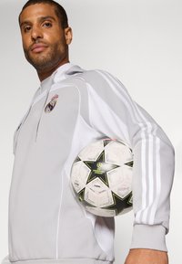 Siva atletska jakna s bijelim prugama i logom Real Madrida, drži bijelu nogometnu loptu s dizajnom zelenih i crnih zvijezda.