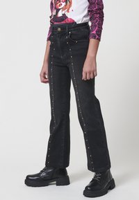 Jeans negros de cintura alta con tachuelas de latón verticales en el frente, combinados con botas negras gruesas con cordones y una camiseta gráfica de manga larga.
