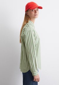 Maglietta a maniche lunghe a righe verdi e bianche con colletto, abbinata a un cappello rosso. La maglietta ha una vestibilità comoda e polsini con bottoni.