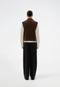 Brązowa kamizelka z korduroy na kremowym swetrze, zestawiona z luźnymi czarnymi spodniami. Projekt jest minimalistyczny, bez widocznych elementów metalowych.