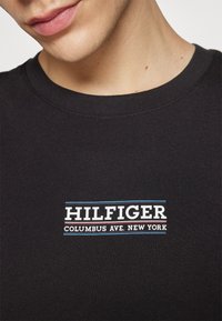 Černé bavlněné tričko s bílým grafickým logem, na kterém je napsáno "HILFIGER" a "COLUMBUS AVE. NEW YORK" s modrými a červenými akcenty. Měkký materiál.