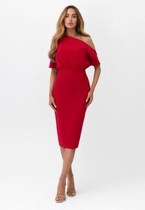 Kobieta o długich włosach w dopasowanej czerwonej sukience midi z jednym rękawem na ramię oraz beżowych sandałach na wysokim obcasie, stojąca na tle białego tła.