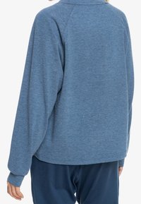 Modrý fleecový pulovr se strukturovaným povrchem, uvolněným střihem a dlouhými rukávy. Obsahuje kulatý výstřih a kleslé švy na ramenou.