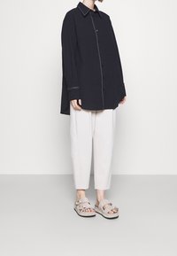 Chemise noire à boutons avec surpiqûres blanches, coupe ample ; pantalon beige clair fuselé ; sandales beiges avec boucles et matériau texturé.