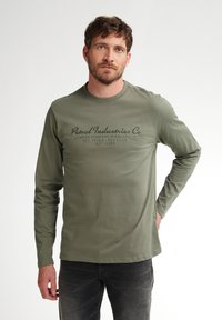 Camică din bumbac de culoare verde măsliniu, cu mâneci lungi, guler rotund, text imprimat în negru și croială relaxată. Pur tată cu blugi negri.