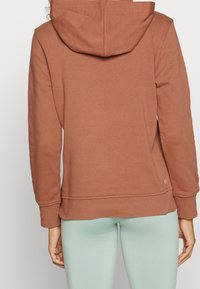 Brun hættejakke med snøre, ribbede mansjetter og lige kant, lavet af blødt stof, båret over lysegrønne leggings.