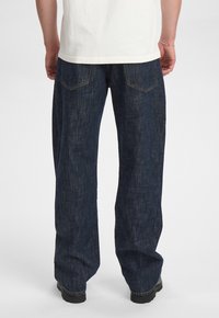 Jeans in denim blu scuro con una vestibilità ampia, caratterizzati da gamba dritta, due tasche posteriori e dettagli di cucitura sottili, su uno sfondo chiaro.
