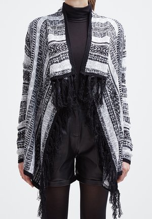 Zwart-wit patroon open cardigan met franjeranden, gedragen over een zwarte coltrui en zwarte leren korte broek.