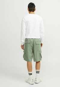 Chemise blanche légère à manches longues associée à un short cargo vert olive, doté de plusieurs poches latérales, portée avec des chaussettes rayées et des baskets.