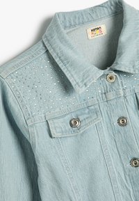 Giacca di jeans blu chiaro con colletto, caratterizzata da un design borchiato sulle spalle, hardware argentato e una tasca sul petto con chiusura a scatto.