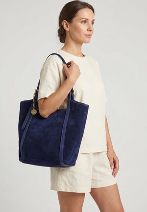 Kvinde iført beige outfit bærer en stor marineblå ruskindstaske med lange stropper over skulderen mod en ensfarvet baggrund.