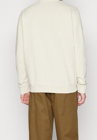 Felpa color beige chiaro con maniche lunghe e polsini a coste. Rifinita con un collo rotondo e un orlo dritto, abbinata a pantaloni marroni.