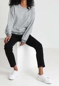 Sudadera gris de cuello redondo con banda de logo negra, combinada con jeans negros y zapatillas blancas. La tela parece suave y tiene un corte relajado.