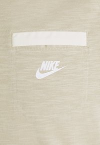 Jasno beżowa tkanina o teksturowym wykończeniu, z białą naszywaną kieszenią i białym logo Nike wydrukowanym w dolnym prawym rogu.