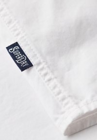 Witte stof met een gladde textuur, met een gevouwen hoek en een donkerblauw label met "Superdry" in witte stiksel.