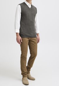 Grauer gestrickter ärmelloser Pullover mit V-Ausschnitt, getragen über einem weißen Hemd mit Kragen. Kombiniert mit hellbraunen Slim-Fit-Hosen und beigen Stiefeletten.