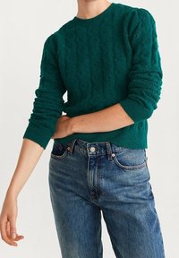Pull vert en maille avec motif texturé, poignets côtelés et col rond, associé à un jean denim bleu.