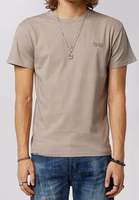 T-shirt in cotone color taupe chiaro con collo rotondo, maniche corte e piccolo logo ricamato sul petto. Abbinato a jeans in denim strappati.