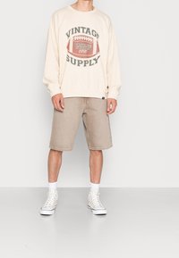 Osoba oblečená v béžovém vintage svetru s fotbalovou grafikou, khaki kraťase po kolena, bílých ponožkách a bílých vysokých teniskách.