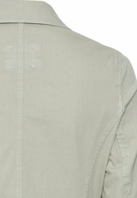 Partie arrière d'une veste beige clair avec des coutures visibles le long des coutures centrales et des épaules.