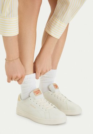 Persona con zapatillas blancas de Reebok y calcetines blancos, ajustándose los calcetines, con las mangas de una camisa con rayas amarillas remangadas visibles.