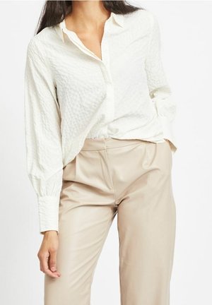 Kobieta ubrana w białą koszulę z fakturą, zapinaną na guziki, z długimi rękawami oraz beżowe spodnie z wysokim stanem, z ręką opartą wzdłuż ciała.