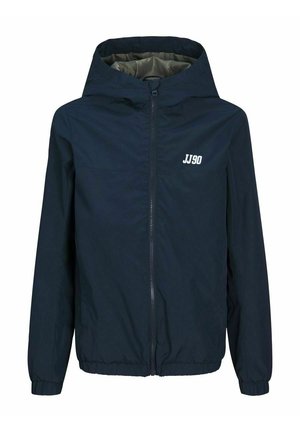 Enfant portant une veste bleu marine, une chemise vert olive, un jean skinny gris et des baskets montantes noires avec des lacets blancs, debout devant un fond uni.