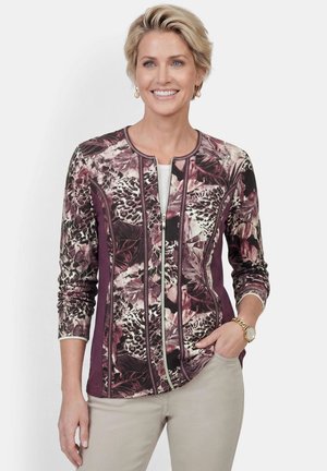Femme souriante aux cheveux blonds courts porte une veste zippée violette à motifs floraux et animaux et un pantalon beige, avec une montre et des boucles d'oreilles.