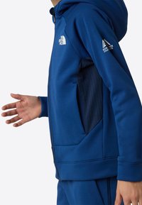 Kék kapucnis pulóver puha anyagból, cipzárral, oldalzsebekkel és kontrasztos, texturált panelekkel. Jelvények a mellkason és az ujján.