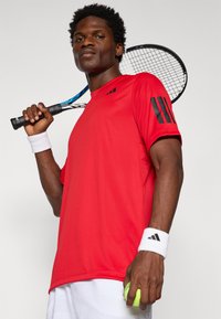 Camiseta deportiva roja con acentos negros, muñequeras blancas, y sosteniendo una raqueta de tenis. El modelo tiene el cabello corto y rizado y está en una pose confiada.