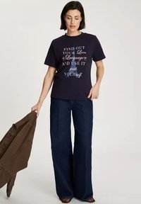 Granatowa koszulka z napisem "ODKRYJ SWÓJ JĘZYK MIŁOŚCI I UŻYWAJ GO Z SOBĄ" w białym i szarym kolorze. Połączona z szerokimi dżinsami i brązową kurtką.