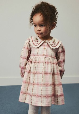 Junges Mädchen mit lockigem Haar, das ein langärmliges rosa kariertes Kleid mit Blumenstickerei am Kragen trägt, steht auf einem blauen Teppich.