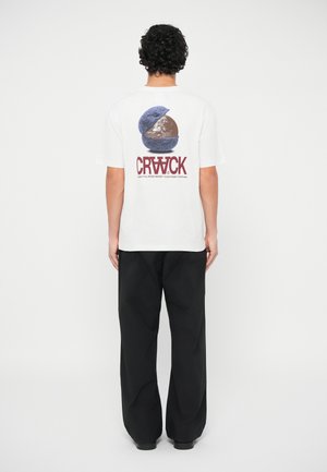 Λευκό βαμβακερό t-shirt με γραφικό σχέδιο ενός διαιρεμένου γηπέδου και το κείμενο "CRWCK." Συνδυάζεται με μαύρα φαρδιά παντελόνια.