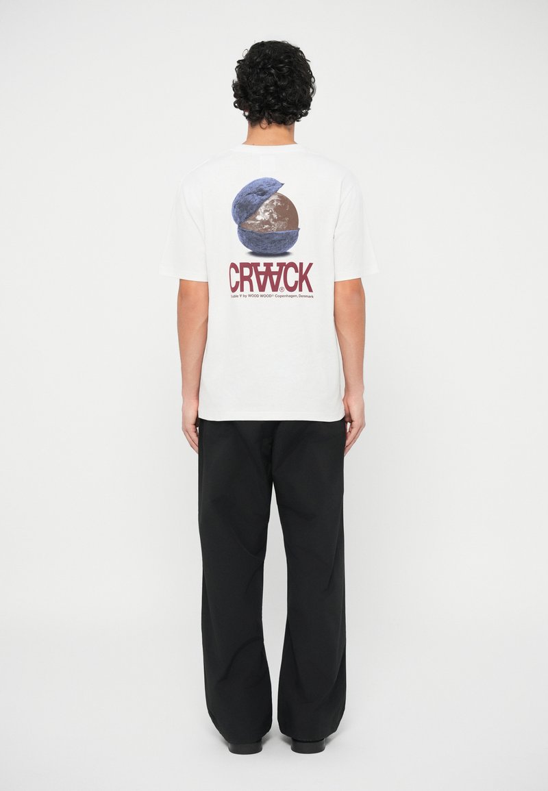 Λευκό βαμβακερό t-shirt με γραφικό σχέδιο ενός διαιρεμένου γηπέδου και το κείμενο "CRWCK." Συνδυάζεται με μαύρα φαρδιά παντελόνια.