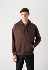 Young man wearing a brown zip-up hoodie with drawstrings and front pockets, paired with dark jeans, standing against a plain light background.