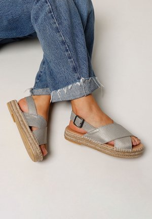 Sølvfarvede sandaler med krydsede remme i tekstureret læder, en flettet jutesål og en justerbar ankelrem, kombineret med slidte blå jeans.