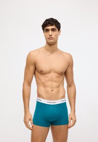Tirkīza kokvilnas bokseršortiem ar baltu elastīgu jostasvietu, uz kuras atrodas "Calvin Klein" logo, pieguļošs dizains un gluda tekstūra.