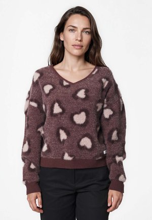 Femme portant un pull marron duveteux avec des motifs de cœurs rose clair et un pantalon noir, debout sur un fond blanc uni.