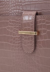 Vaaleanruskea tekokrokotiilinnahkainen laukku, jossa on ommeltu hihna ja kapea vaakasuora kultainen metallilukko.