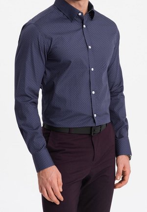 Homme portant une chemise à manches longues ajustée, bleu foncé à motifs, boutonnée et rentrée dans un pantalon violet foncé avec une ceinture noire.