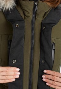 Olivgrön isolerad jacka med svarta detaljer, som har en dragkedja framtill, tryckknappar och en krage av konstpäls. Strukturerad tyg.